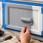 Close up of renter friendly kitchen cabinets being painted soft grey with a foam roller, blue painter’s tape protecting edges and a drop cloth covering the floor to show careful, non permanent renovation.