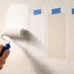 Close up of a hand using a paint roller to apply fresh white paint on a textured wall, showing warm white and cool white paint swatches side by side with blue painter’s tape labels and natural side lighting.